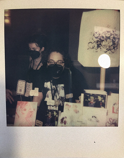 a scanned polaroid of the site owner, a white person with frizzy brown hair and glasses wearing a black n95 mask, behind a table at the comic show SPX